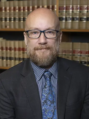 Man wearing glasses with beard in suit and blue tie in front of shelves of law books